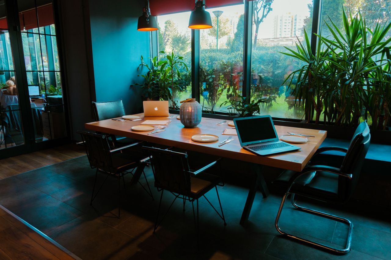 about-us A modern dining room setup with laptops, plates, and lush plants by large windows.