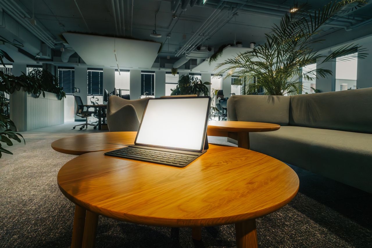 who-we-are Empty office lounge featuring a glowing laptop on a wooden table, surrounded by greenery.